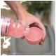 Une main verse de la litière pour chat en cristal rose d'une bouteille dans une tasse à mesurer rose, avec une boîte à litière floue en arrière-plan.