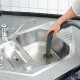 Eine Person verwendet einen Staubsaugerschlauch, um ein Edelstahlspülbecken mit Wasser zu reinigen, ausgestattet mit einem modernen Wasserhahn und einer grauen Arbeitsplatte.