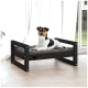 Ein Hund, der auf einem stilvollen schwarzen Holz-Haustierbett mit einem grauen Kissen ruht, in einem hellen Raum mit großen Fenstern und einer Pflanze in der Nähe.