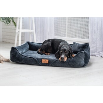 Schwarz-brauner Hund liegt auf einem dunkelgrauen Plüsch-Haustierbett mit "tierlando"-Etikett in einem hellen Raum.