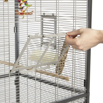 Eine Hand einer Person öffnet eine metallene Zugangstür zu einem Haustierkäfig und enthüllt eine Holzstange und einen hängenden Leckerli-Stick im Inneren.