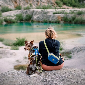 Eine Frau mit kurzen blonden Haaren sitzt auf einem felsigen Vorsprung am türkisfarbenen Wasser, während ihr braun-weißer Hund in die Kamera schaut.