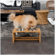 Ein flauschiger Pomeranian-Hund frisst aus einer stilvollen Bambus-Futterstation mit zwei Edelstahlschüsseln, die in einem modernen Wohnzimmer steht.
