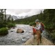 Eine Person mit einem Rucksack sitzt auf einem Felsen am Fluss und blickt auf die Berge, während ein brauner Hund neben ihnen steht.