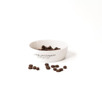 Eine weiße Haustierfutterschüssel mit dem Text "LIEBLINGSNAPF" enthält dunkelbraunes Kibble, mit einigen verstreuten Stückchen darum.