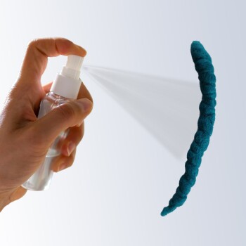 Eine Hand hält eine Sprühflasche, die ein türkisfarbenes Haustierspielzeug in Wurmform gegen einen hellen Hintergrund besprüht.