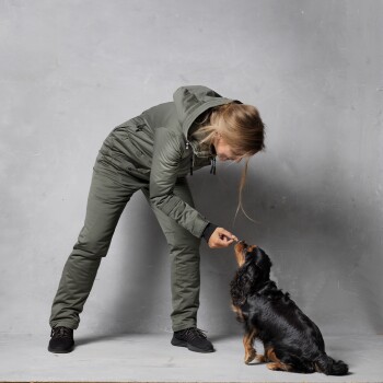 Eine Person in einer grünen Jacke und Hose füttert einen kleinen schwarz-braunen Hund mit einem Leckerli, vor einem grauen Hintergrund.