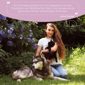 Eine Frau mit einer schwarzen Katze sitzt neben einem wolfähnlichen Hund in einem Garten mit lila Blumen. Der Text darüber bewirbt Wolfsbacher Naturprodukte.