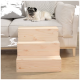 Ein Mops-Hund ruht auf einem hellgrauen Sofa mit einem gemusterten Kissen, neben einer hölzernen dreistufigen Haustiertreppe, die für einen einfachen Zugang konzipiert ist.