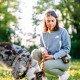 Eine Frau in einem grauen Sweatshirt und grünen Hosen kniet im Gras, hält eine Leine, während ihr Hund im gemusterten Geschirr den Boden schnüffelt.