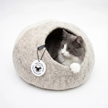 Eine Siamkatze mit blauen Augen, die aus einem gemütlichen, grauen Filz-Haustierbett in Form eines Steins schaut, vor einem weißen Hintergrund.