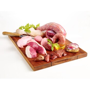 Ein Holzschneidebrett hält rohes Huhn, Schweinefleisch und Innereien, garniert mit Kräutern und einer kleinen Schüssel Öl.