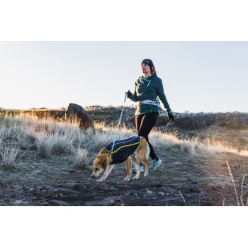 Eine Frau in einer türkisfarbenen Jacke und schwarzen Leggings führt einen Golden Retriever, der einen blauen gemusterten Mantel trägt, auf einem felsigen Weg bei Sonnenuntergang.