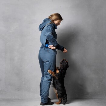 Eine Person in einem blauen Overall steht mit einem kleinen Hund, der zu ihnen hochgreift, möglicherweise nach einem Spielzeug oder Leckerli.