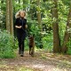 Eine Person in schwarzer Sportkleidung joggt auf einem Waldweg mit einem braunen Hund an der Leine, umgeben von üppigem Grün.