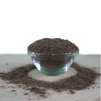 Eine klare Glasschüssel gefüllt mit einem feinen, dunkelbraunen Pulver, umgeben von einem kleinen Haufen des gleichen Pulvers auf einer hellen Oberfläche.