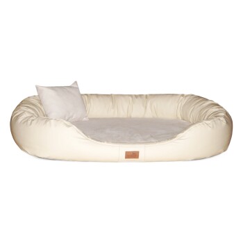 Ein weißer Hund, der auf einem cremefarbenen Haustierbett mit einem kleinen Kissen entspannt, vor einer neutralen Wand und einer Pflanze in einem Topf.