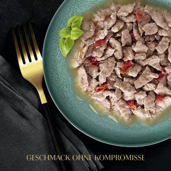 Nahaufnahme eines grünen Tellers mit gehacktem Haustierfutter, Basilikumblättern und getrockneten Tomaten, mit dem Text "GESCHMACK OHNE KOMPROMISSE."