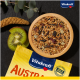 Eine Holzschale mit buntem Vogelfutter steht auf einer dunklen Oberfläche mit einer geschnittenen Kiwi und einem gelben Vitakraft-Paket beschriftet "AUSTRALIA."