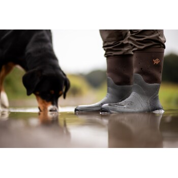 Person trägt braune und graue wasserdichte Stiefel und steht in einer Pfütze, während ein schwarz-brauner Hund in der Nähe Wasser trinkt.