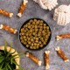 Eine schwarze Schüssel, gefüllt mit kleinen, grünlich-braunen Hundefutter-Kibble, umgeben von Hundekau-Knochen und einer Pflanze, auf einer grauen Oberfläche.