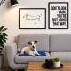 Ein kleiner Hund liegt auf einer blauen Matte auf einem grauen Sofa, mit zwei gerahmten Kunstwerken an der Wand: einer einfachen Strichzeichnung eines Hundes und einem motivierenden Zitat.