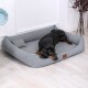 Ein schwarzer und brauner Dobermann liegt bequem auf einem grauen Hundebett mit einem kleinen Kissen, vor einem hellen Holzboden und weißen Wänden.