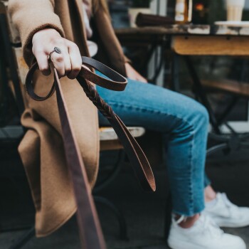 Eine Person hält eine braune Leder-Hundeleine, sitzt auf einem Stuhl in einem Café. Sie trägt einen beigen Mantel und blaue Jeans, mit weißen Sneakers.