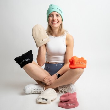 Eine Frau in einem weißen Tanktop und blauen Shorts sitzt im Schneidersitz, hält eine beige Mütze und ist von bunten "HUFIESTER" Mützen umgeben.