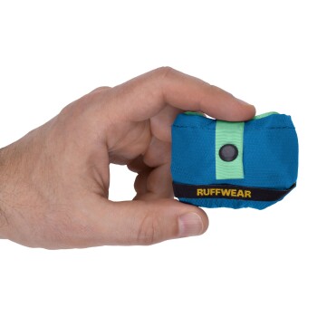 Eine Hand hält eine kleine, blaue Haustier-Zubehörtasche mit einem grünen Riemen und einem schwarzen Knopf, beschriftet mit "RUFFWEAR."