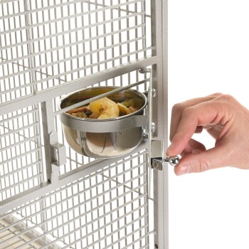 Eine Hand sichert eine Metallfutterschüssel, die an einem Haustierkäfig befestigt ist, gefüllt mit buntem Haustierfutter und getrockneten Obstscheiben.