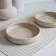 Zwei beige Keramik-Haustiernäpfe stehen auf einer weißen gestreiften Matte, begleitet von einer strukturierten beigen Vase und einer schlichten weißen Vase im Hintergrund.