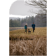Eine Frau in einer türkisfarbenen Jacke und ein Mann in einer blauen Jacke führen einen schwarzen Hund auf einem grasbewachsenen Weg in der Nähe eines Teiches, umgeben von Bäumen.
