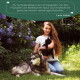 Eine Frau im Freien mit Blumen hält eine schwarze Katze, während ein Siberian Husky in der Nähe ruht. Ein Zitat von Lena Huber über Wolfsbacher Natur steht darüber.