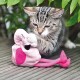 Ein verspieltes graues Tabby-Kätzchen, das mit seinen Pfoten ein weiches pinkes Flamingo-Spielzeug greift, umgeben von grünem Laub auf einer Holzoberfläche.