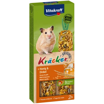 Uroczy beżowy chomik siedzi obok pudełka Vitakraft Kräcker z przysmakami miodowymi i orkiszowymi, oznaczonymi jako bogate w witaminy i bez cukru.
