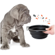 Ein grauer Welpe mit faltiger Haut sitzt neben einer zusammenklappbaren schwarzen Fressnapf, die von einer Hand gehalten wird, mit bunten Spritzern, die in der Nähe illustriert sind.
