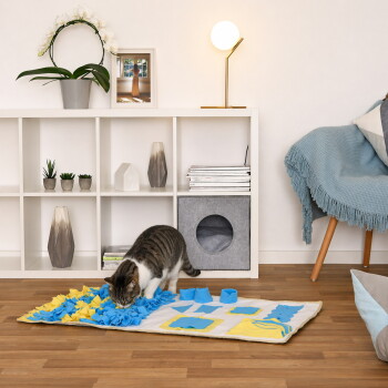 Getigerte Katze schnüffelt an einer blau-gelben Snuffle-Matte auf dem Boden in einem modernen Wohnzimmer mit weißem Würfelregal und grauem Katzenwürfel.