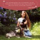 Eine Frau mit langen Haaren hält eine schwarze Katze neben einem wolfähnlichen Hund in einem Garten mit lila Blumen. Text oben bewirbt Wolfsbacher Natur.