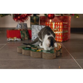 Tabby and white cat playing on a Christmas tree-shaped cardboard scratcher with wrapped gifts in the background.