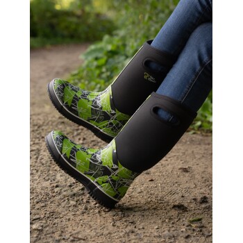 Ein Paar stilvolle grün-schwarz gemusterte Gummistiefel, die von einer Person auf einem Kiesweg getragen werden, umgeben von Grün.
