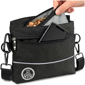 Eine Hand, die ein Hundeleckerli in eine schwarze Hundeleckerli-Tasche mit Reißverschluss und einem Logo steckt, mit einem Schultergurt und reflektierenden Details.