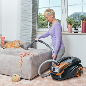 Eine Frau in einem lila Pullover saugt ein Sofa mit einem schwarz-orangen Staubsauger, während eine rote Katze zuschaut. Garn ist auf dem Sofa verstreut.