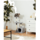 Eine getigerte Katze sitzt auf einem grauen Katzenbaum mit zwei Plattformen und einem Kratzbaum, neben einer weißen Kommode und einer blättrigen Pflanze.