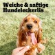 Ein glücklicher golden Cocker Spaniel schaut gespannt auf eine Hand, die ein Leckerli anbietet, mit "Weiche & saftige Hundeleckerlis" darüber.