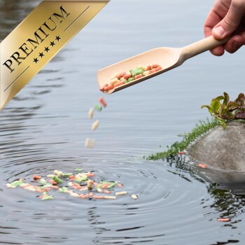 Eine Hand, die farbenfrohes Fischfutter aus einer Holzschaufel in einen Teich gießt, wobei Wellen im Wasser entstehen. Ein "PREMIUM"-Goldlabel befindet sich in der Ecke.