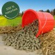 Umgekippter roter Eimer verschüttet braune Tierfutterpellets auf Stroh, grüner Feldhintergrund; rundes Siegel: '100% natürlich'