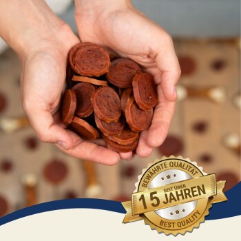 Hände halten einen Stapel runder, rötlich-brauner Haustierleckereien, mit einem Abzeichen, das 'Beste Qualität' und '15 Jahre' auf Deutsch angibt.
