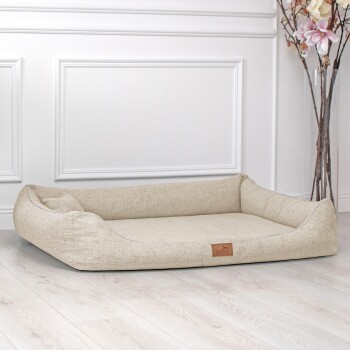 Ein beiges Haustierbett mit erhöhten Rändern und einer kleinen Kopfstütze, mit einem Lederetikett, das 'Pet Landa' liest, auf einem hellen Holzboden platziert.