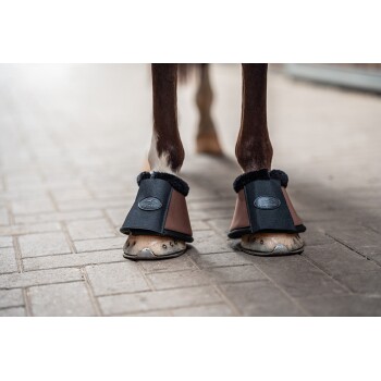 Die Vorderbeine eines Pferdes tragen braune und schwarze Hufschuhe mit einem flauschigen Innenfutter und stehen auf einer strukturierten Ziegeloberfläche.